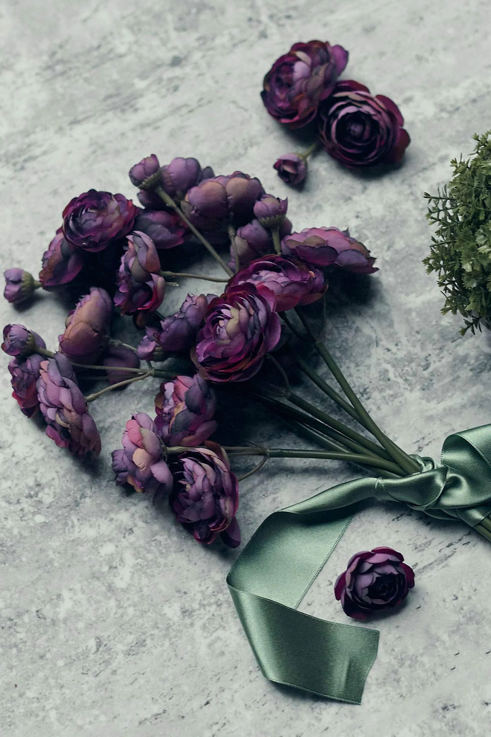 The Abigail Ahern Ranunculus Bunch Purple, featuring vibrant purple blooms with green stems and a green satin ribbon, rests on a gray stone surface, with extra artificial flowers and greenery scattered nearby.