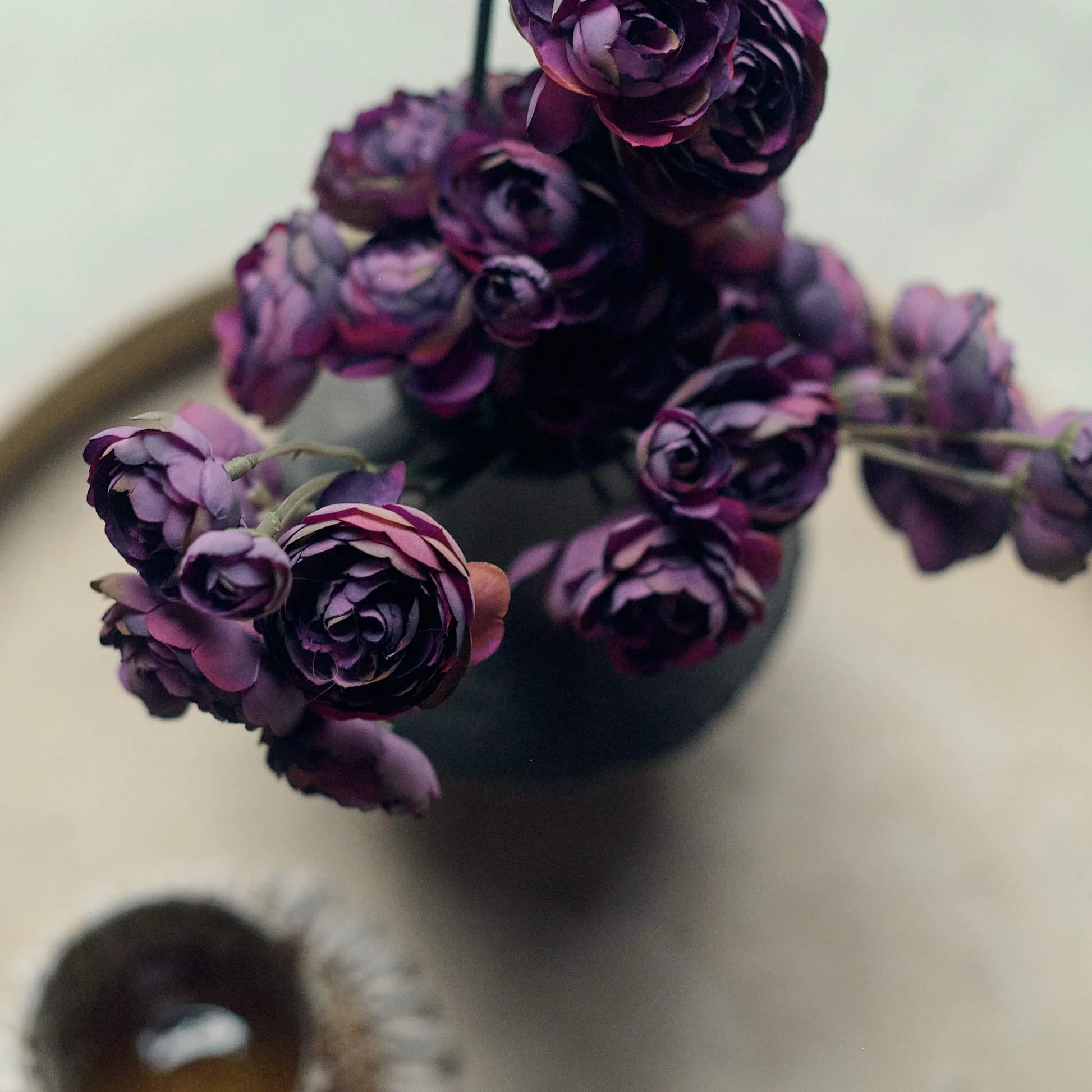 A close-up of the Abigail Ahern Ranunculus Bunch Purple artificial flowers arranged in a vase and viewed from above, with a blurred cup or bowl nearby on a light, round surface.