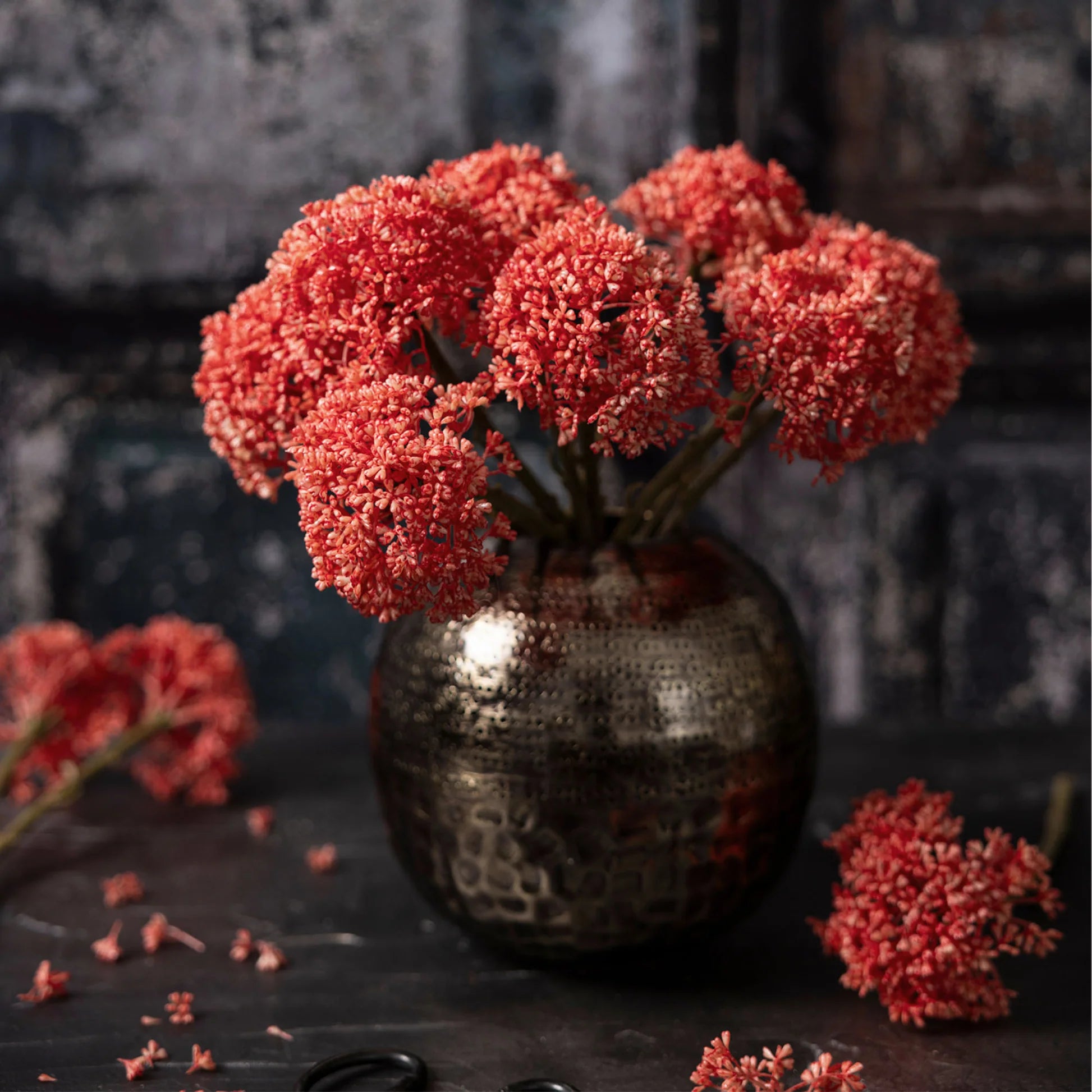 The Abigail Ahern Sedum Coral features vibrant red-orange artificial flowers with clustered petals, arranged in a round, bronze-colored vase on a dark surface, with scattered blooms and scissors against a moody, textured backdrop.