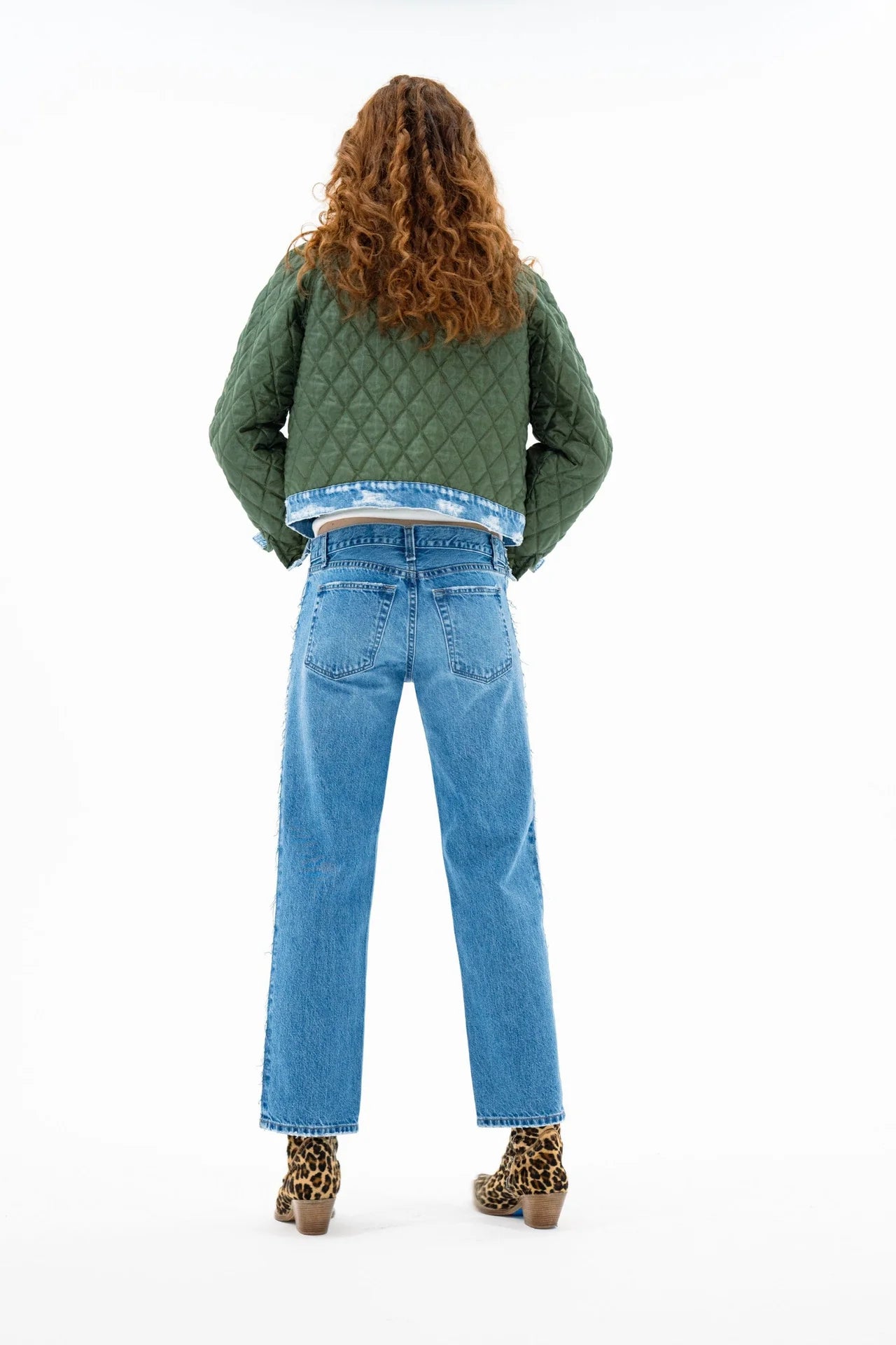 A person with long curly hair faces away, wearing the ASKK Bleeker Street Liner—a green quilted jacket with denim trim—alongside blue jeans and leopard print ankle boots against a white background.