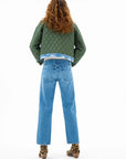 A person with long curly hair faces away, wearing the ASKK Bleeker Street Liner—a green quilted jacket with denim trim—alongside blue jeans and leopard print ankle boots against a white background.