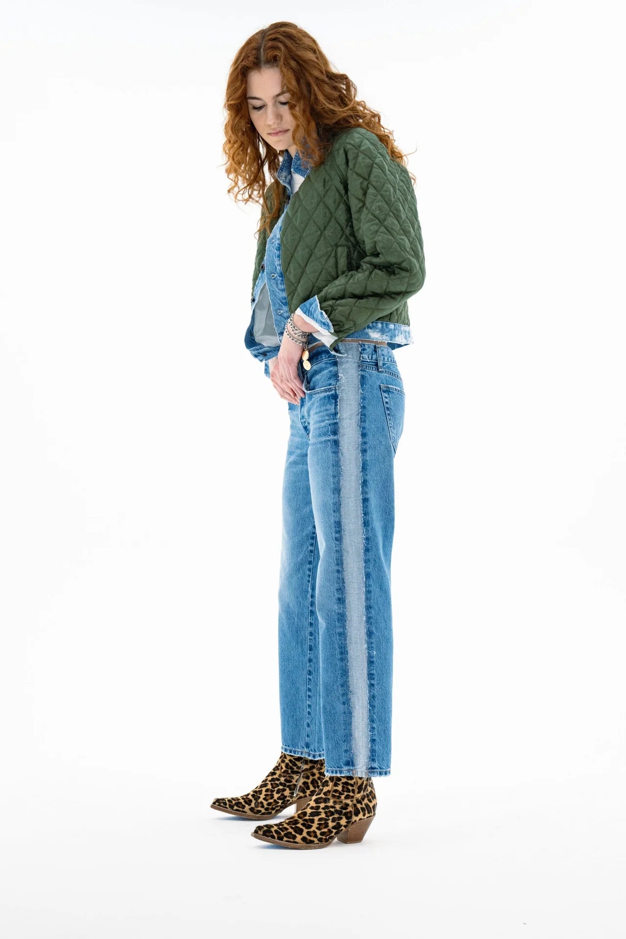 A woman with curly hair wears the ASKK Bleeker Street Liner, a green quilted jacket with denim trim, styled with blue jeans, a light blue shirt, and leopard print ankle boots. She stands looking down against a white background.