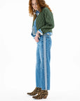 A woman with curly hair wears the ASKK Bleeker Street Liner, a green quilted jacket with denim trim, styled with blue jeans, a light blue shirt, and leopard print ankle boots. She stands looking down against a white background.