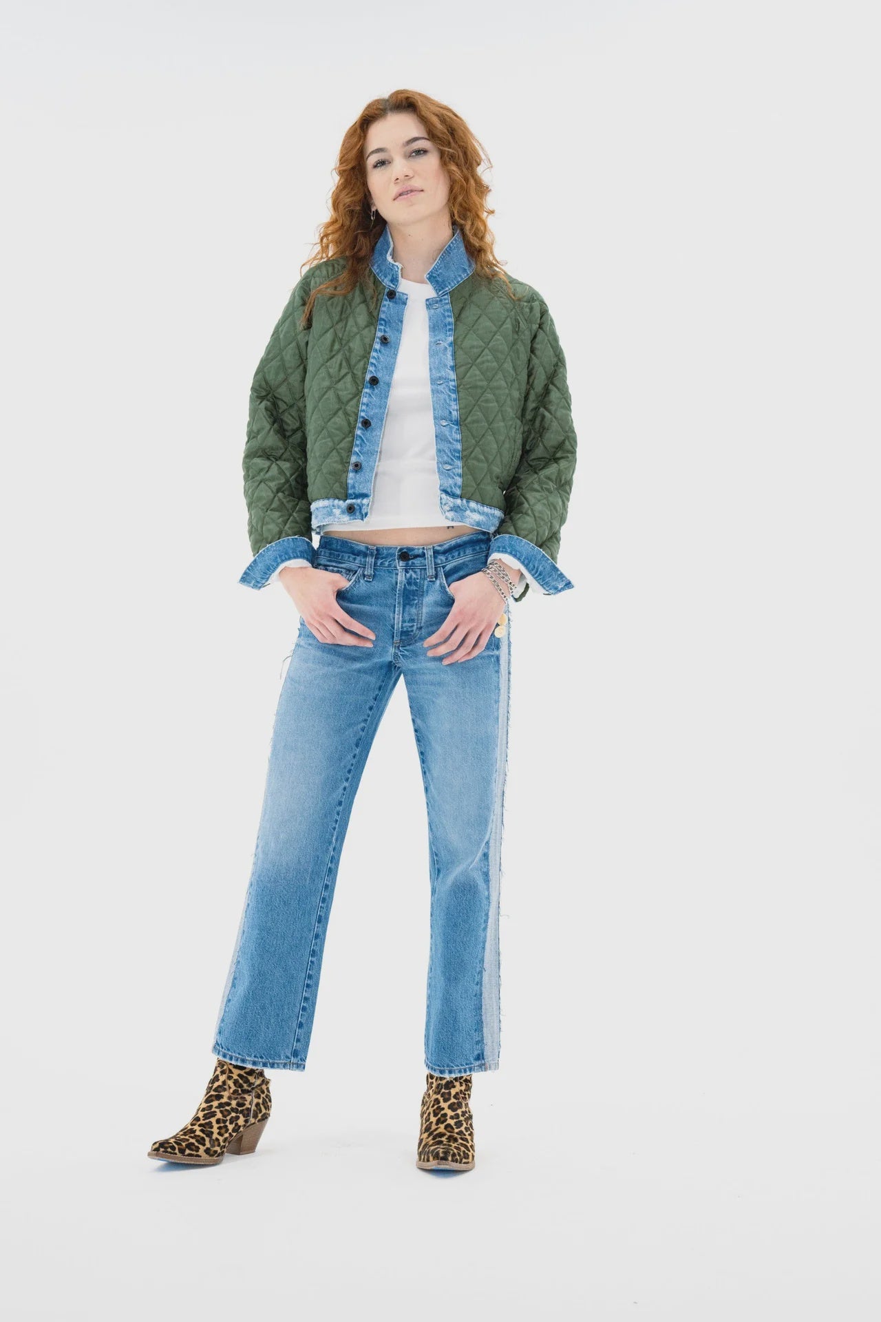 A woman with long red hair models the ASKK Bleeker Street Liner, a green cropped quilted jacket with denim trim, styled over a white shirt and blue jeans with leopard print ankle boots, standing confidently against a plain white background.