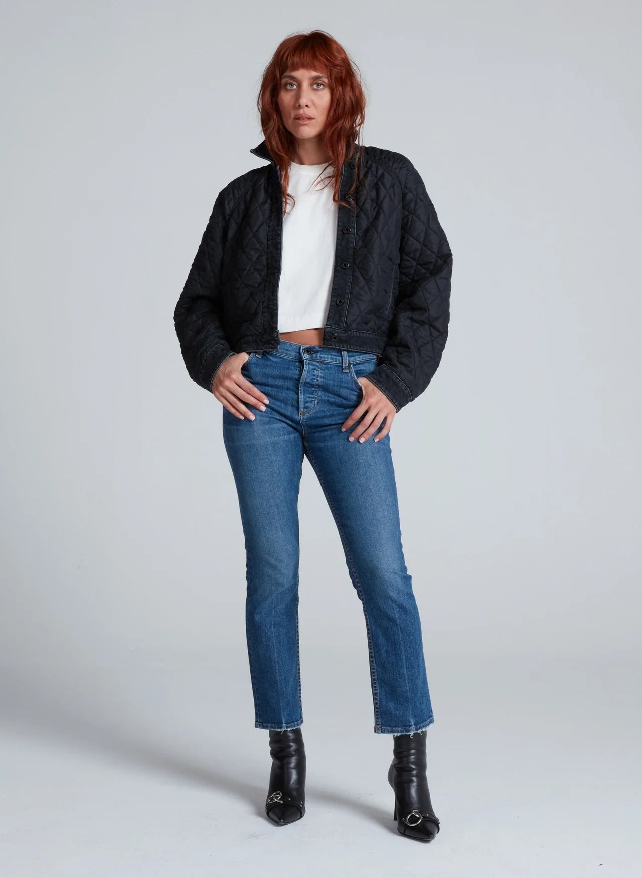 A woman with long reddish-brown hair stands in a studio, wearing the ASKK Bleecker Street Liner—a chic black quilted jacket—over a white crop top, blue jeans, and black heeled ankle boots. She gazes slightly to the side with a neutral expression.
