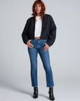 A woman with long reddish-brown hair stands in a studio, wearing the ASKK Bleecker Street Liner—a chic black quilted jacket—over a white crop top, blue jeans, and black heeled ankle boots. She gazes slightly to the side with a neutral expression.