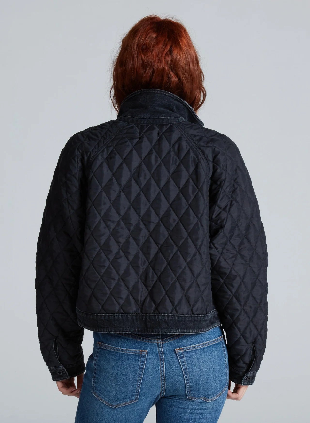 A person with red hair, seen from behind, wears the ASKK Bleecker Street Liner— a black, quilted, cropped jacket—paired with blue jeans against a plain light gray background.