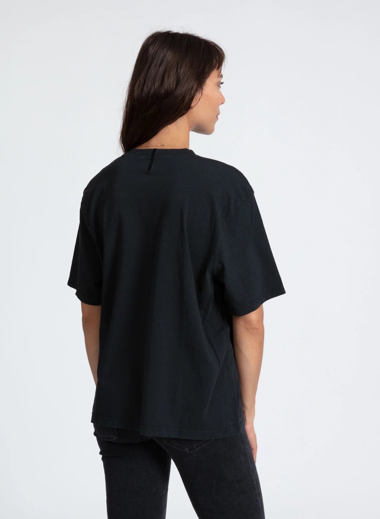 A person with straight brown hair is seen from behind, wearing the ASKK Boy Tee 26, a loose black t-shirt made in Los Angeles, paired with dark jeans against a plain white background.