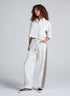 A woman with curly hair wears the ASKK Cancun Pant—white wide-leg drawstring pants with a gray side stripe and elastic waistband—and a cropped white button-up jacket, standing with one hand in her pocket against a plain background.