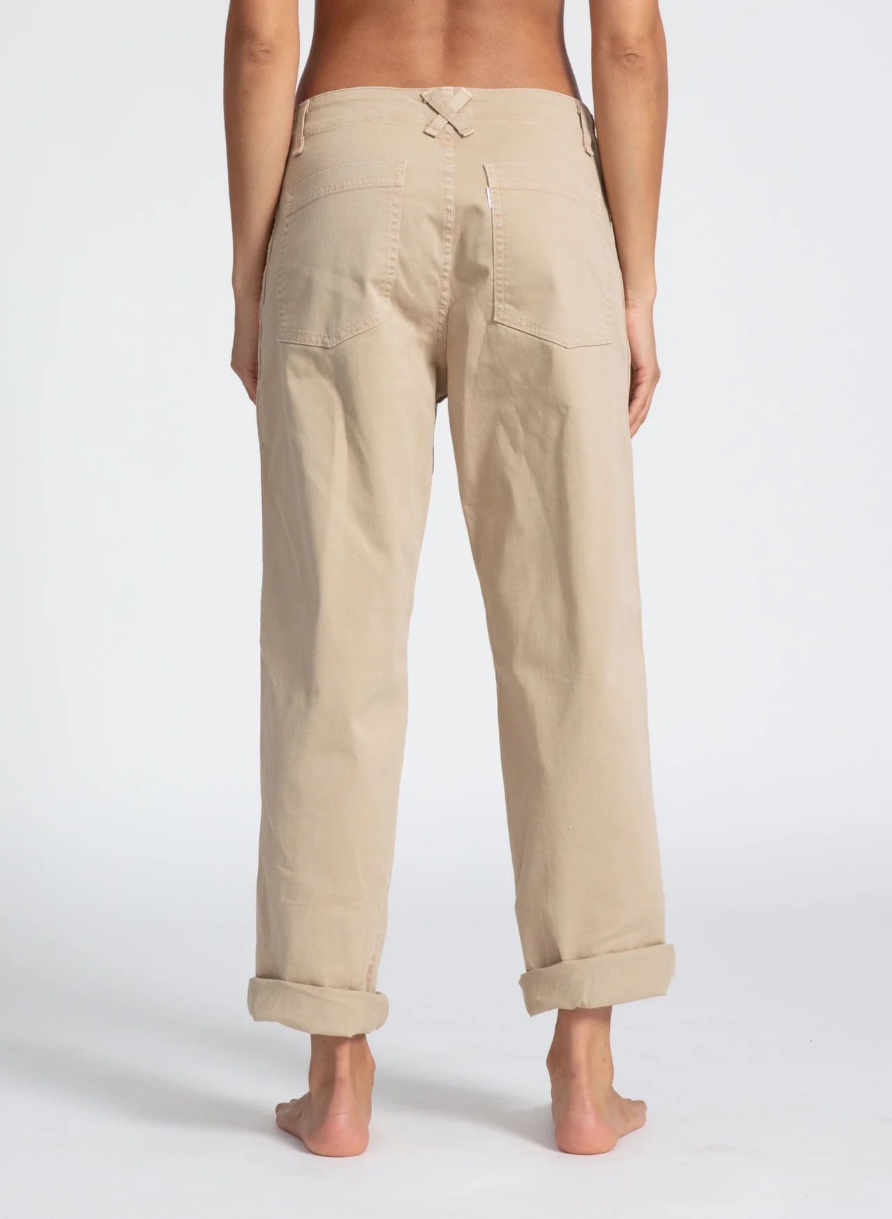 A person stands barefoot facing away, wearing ASKK Slim Chino pants in light beige stretch twill with rolled cuffs and visible back pockets against a plain white background.