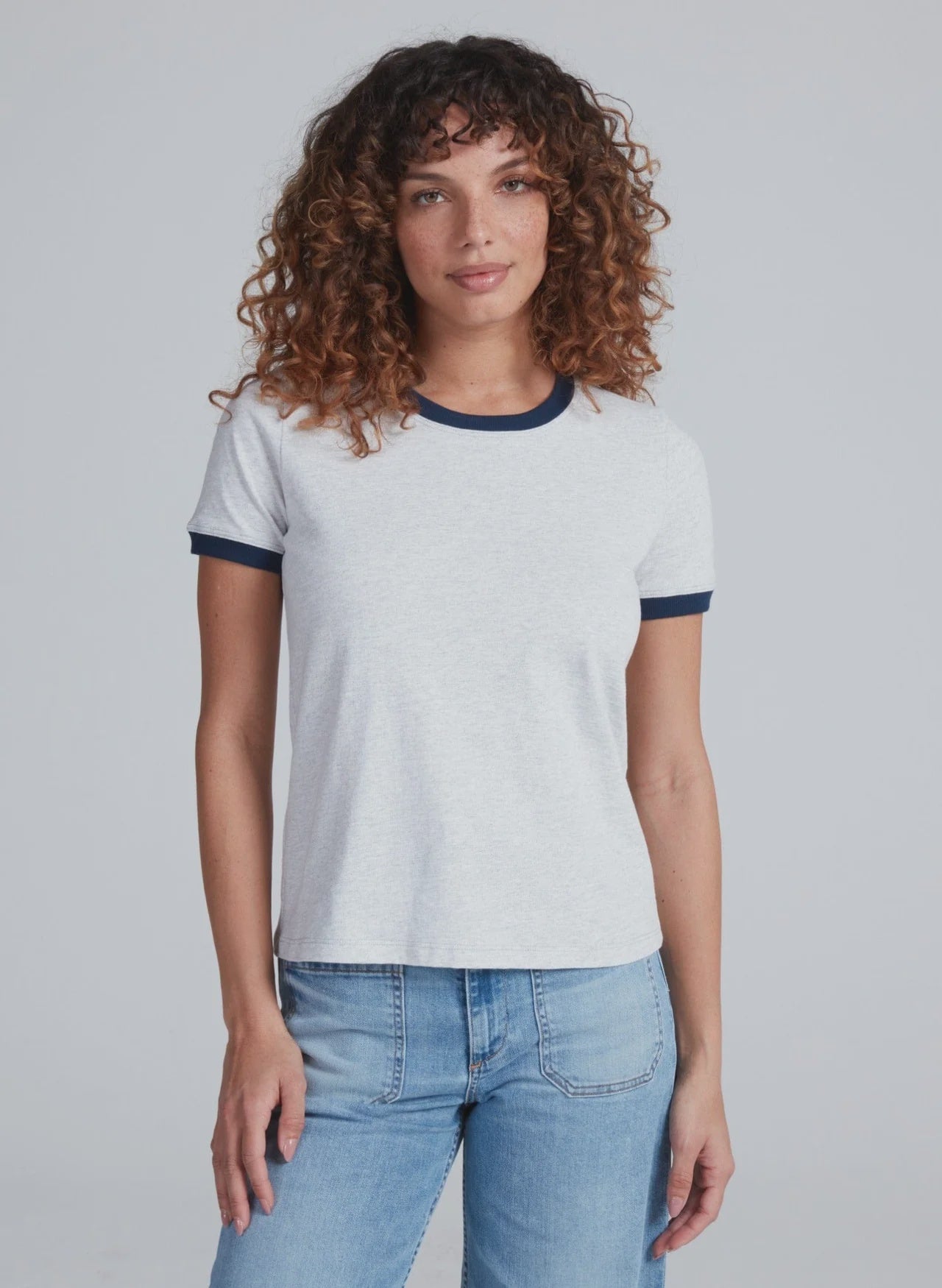 A woman with curly brown hair wears the ASKK Classic Ringer Tee, made from American knit cotton, paired with blue jeans, standing facing forward against a plain light gray background.