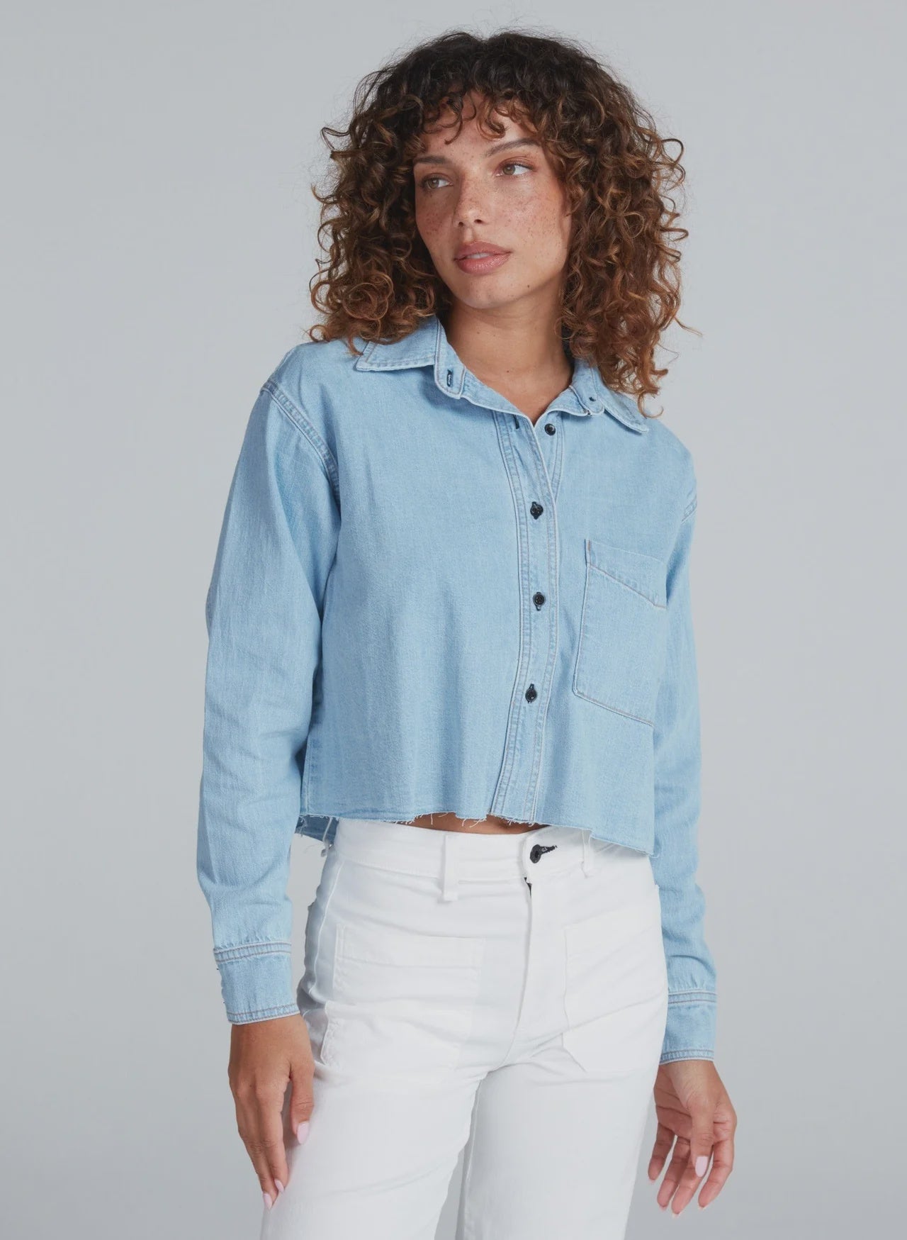 A woman with curly hair models the ASKK Cropped Denim Shirt in light blue with a vintage feel, paired with high-waisted white pants, standing against a plain light gray background.