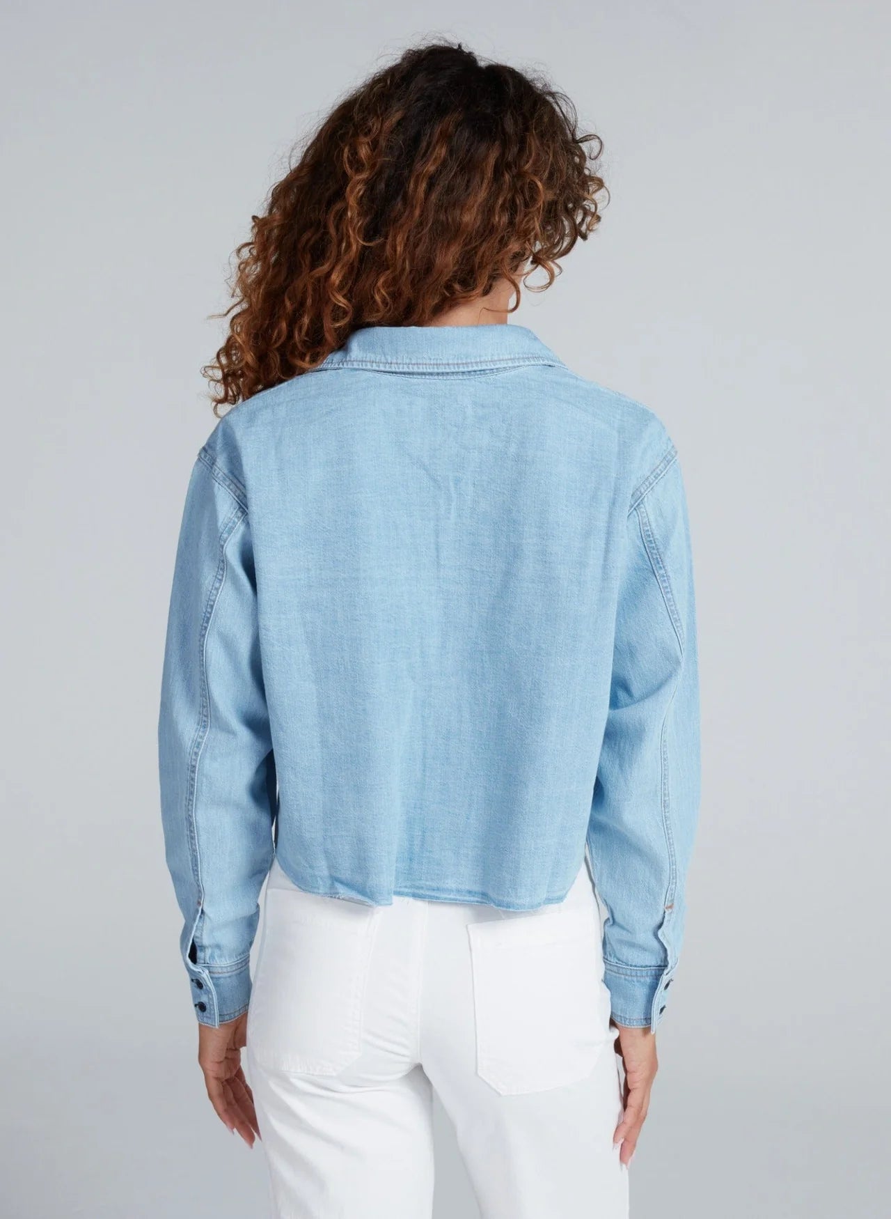 A person with curly hair is seen from the back in an ASKK Cropped Denim Shirt paired with high-waisted white pants, creating a subtle vintage vibe against a plain light gray background.