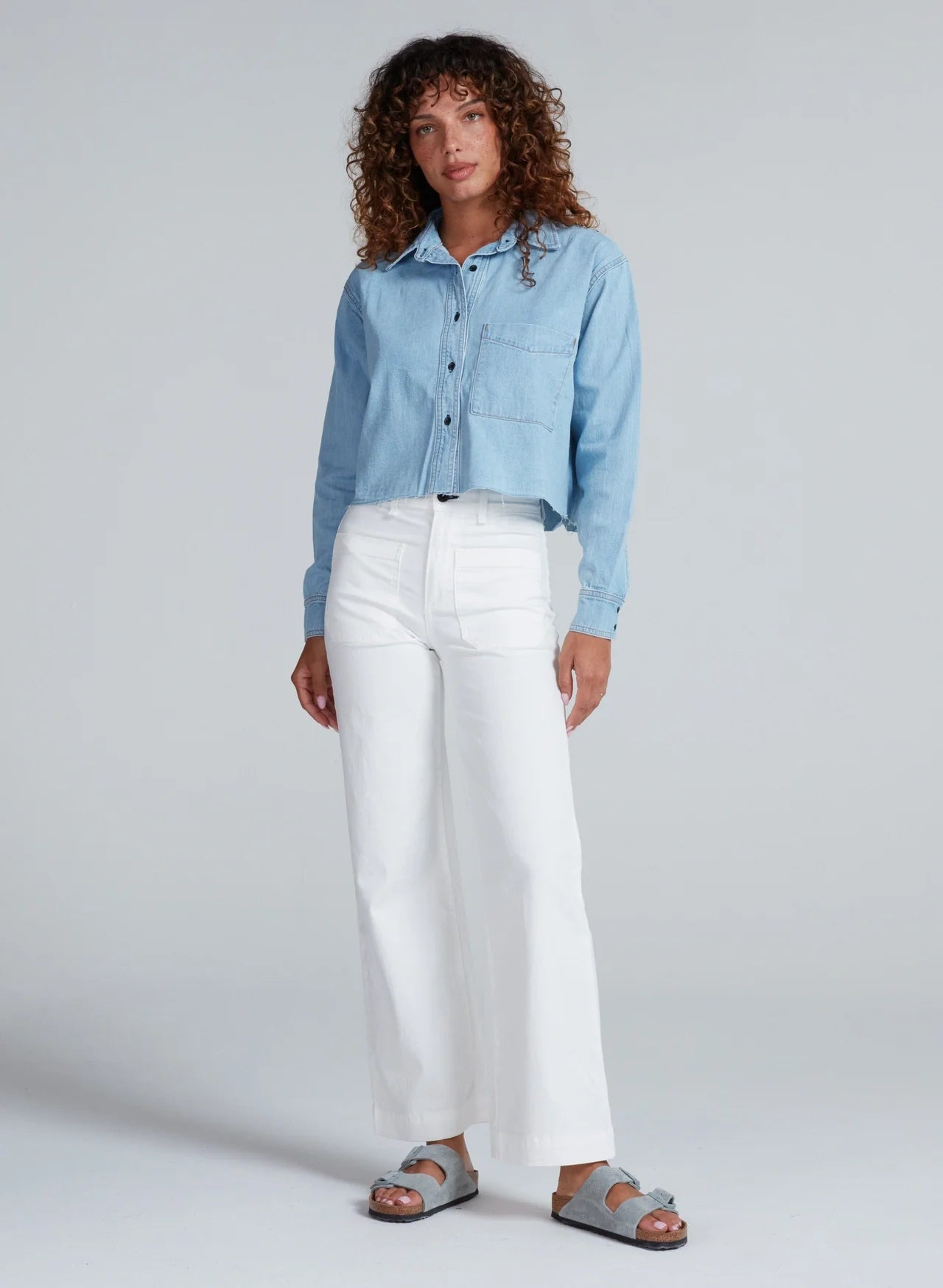 A woman with curly hair wears the ASKK Cropped Denim Shirt in light blue, paired with white high-waisted wide-leg pants and gray sandals. She stands against a plain light gray background, gazing off to the side for a vintage vibe.