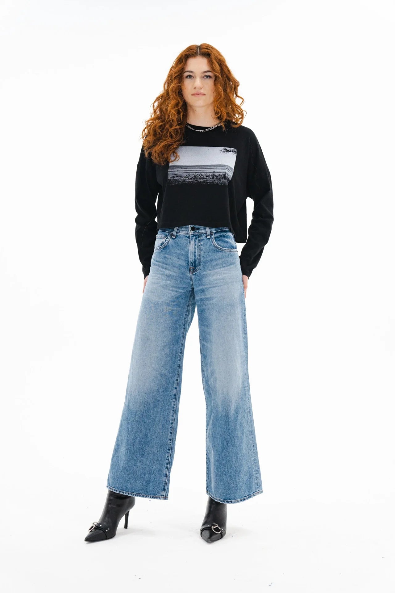 A woman with long curly red hair stands against a white background, wearing an ASKK printed cropped long sleeve shirt, high-waisted wide-leg blue jeans, and black heeled ankle boots.