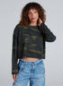 A person with curly brown hair wears the ASKK Cropped LS Tee and light blue jeans, standing against a plain light gray background. One hand touches their hair as they gaze at the camera.
