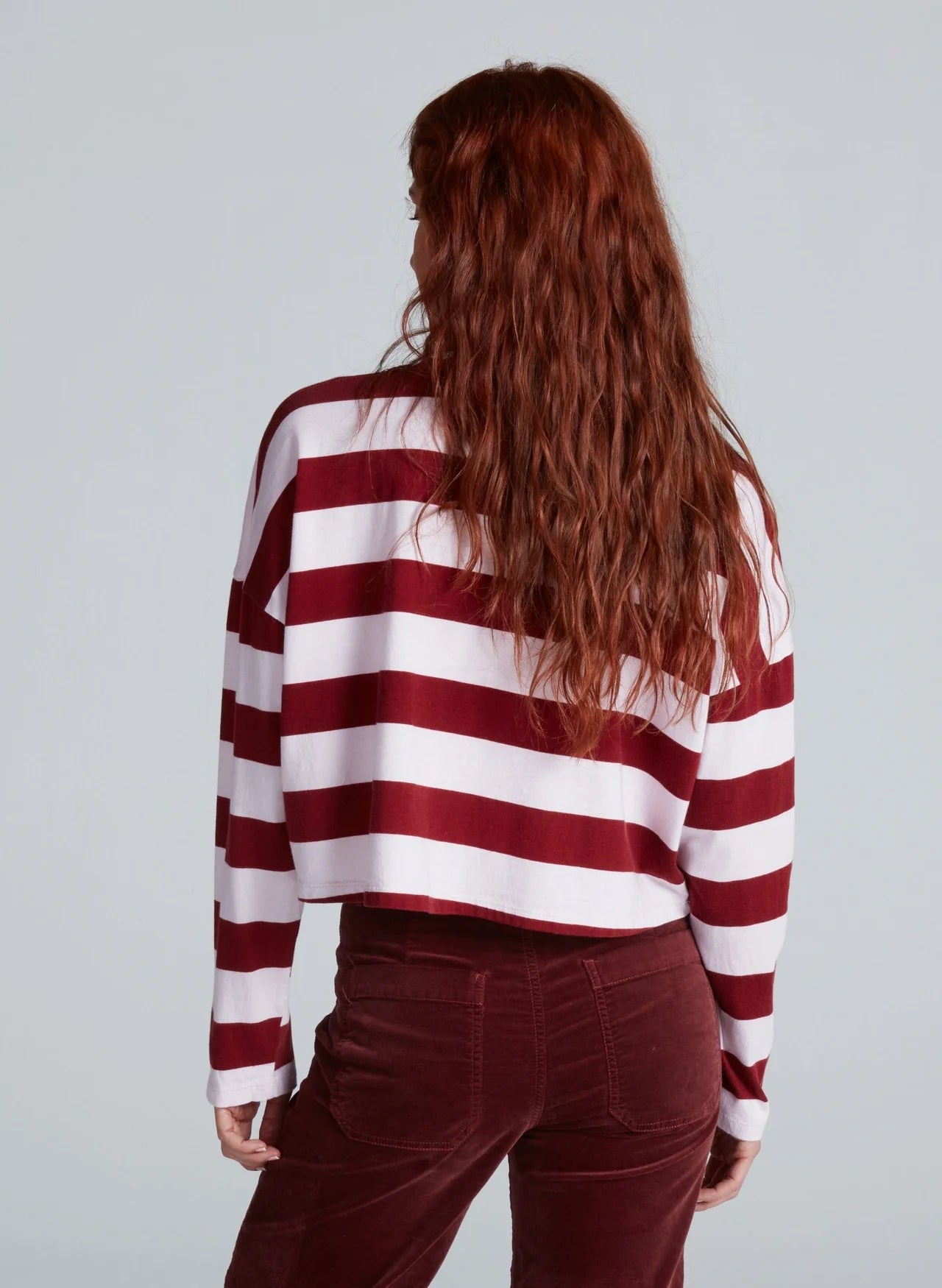 A person with long, wavy auburn hair is seen from behind, wearing the ASKK Cropped LS Tee in red and white stripes and dark red corduroy pants, set against a plain light gray background.