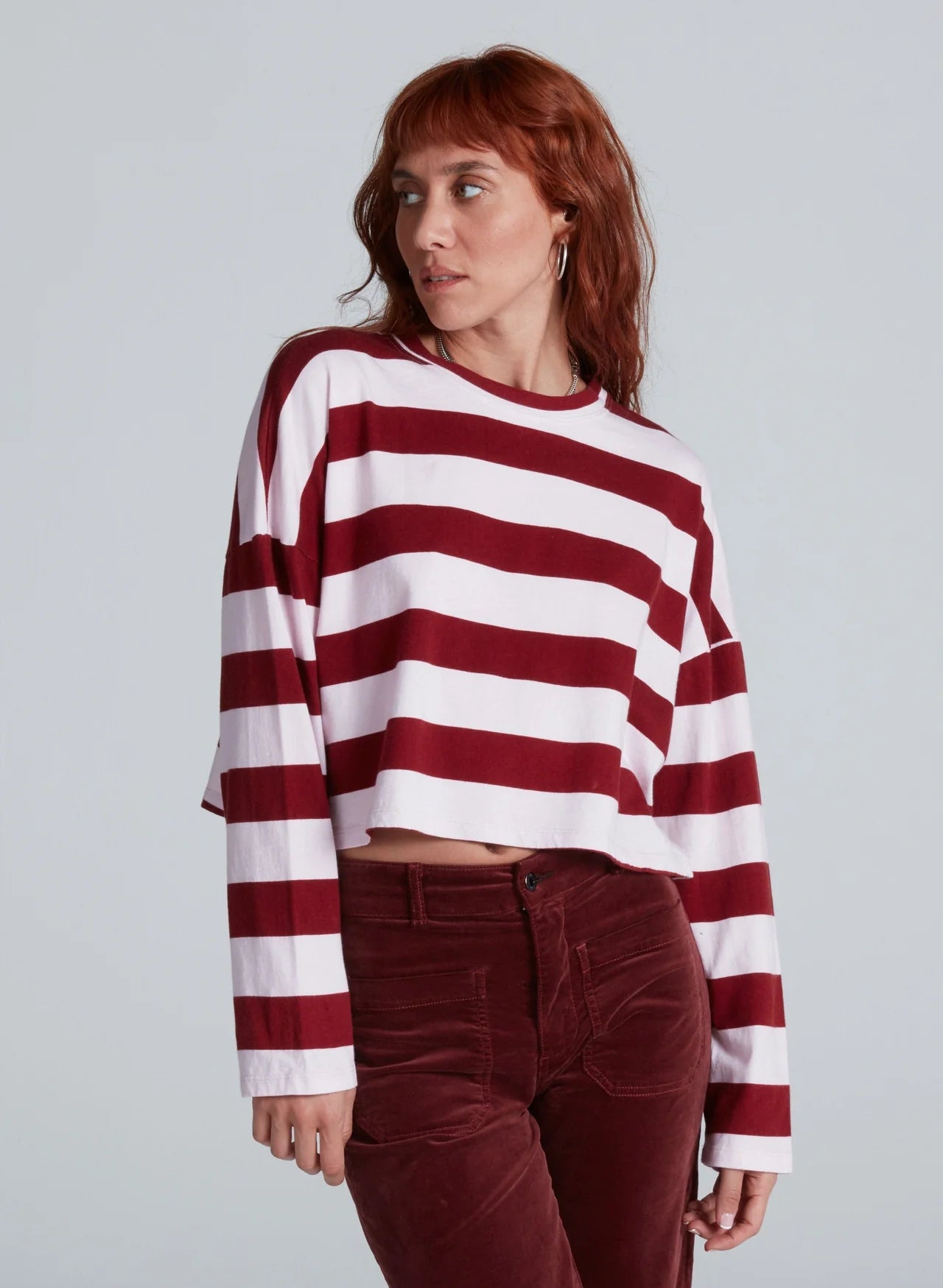 A red-haired person wears the ASKK Cropped LS Tee—long-sleeved, wide maroon and white stripes, soft cotton—paired with high-waisted maroon pants, standing against a plain background.