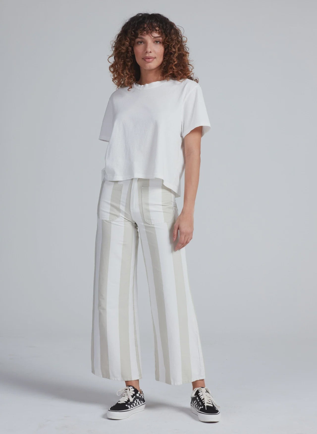 A person with curly hair wears a loose white T-shirt, ASKK’s Cropped Sailor high-rise wide-leg stretch twill pants with beige stripes, and black-and-white sneakers, standing against a plain light gray background.