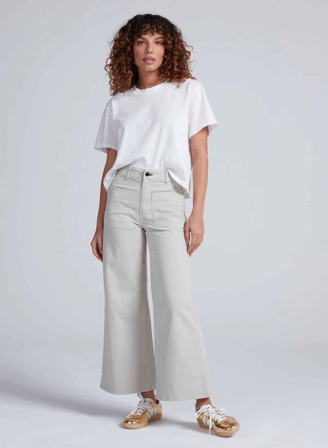 ASKK Cropped Sailor pants in Beach paired with gold sneakers, styled on model with curly hair against gray backdrop.