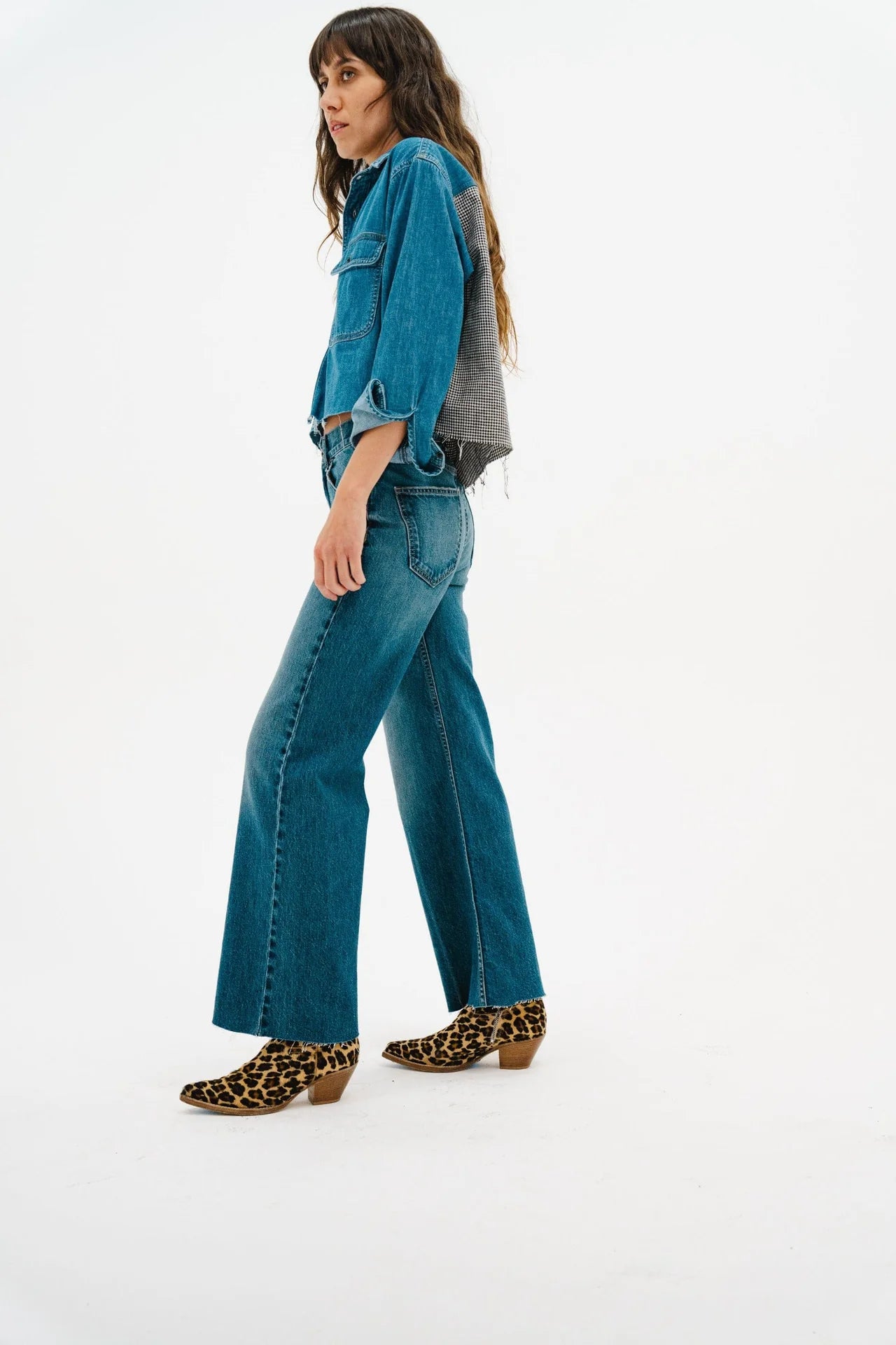A woman with long brown hair stands in profile against a white background wearing an ASKK Crosby Flannel shirt in pink plaid, a blue denim jacket, blue jeans, and leopard-print low-heel ankle boots.