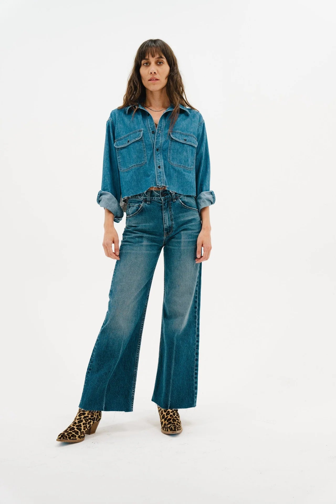 A woman poses against a white background in the ASKK Crosby Flannel, paired with rolled-up sleeves, high-waisted wide-leg jeans, and leopard print ankle boots. Her long brown hair frames her face, completing her chic look.