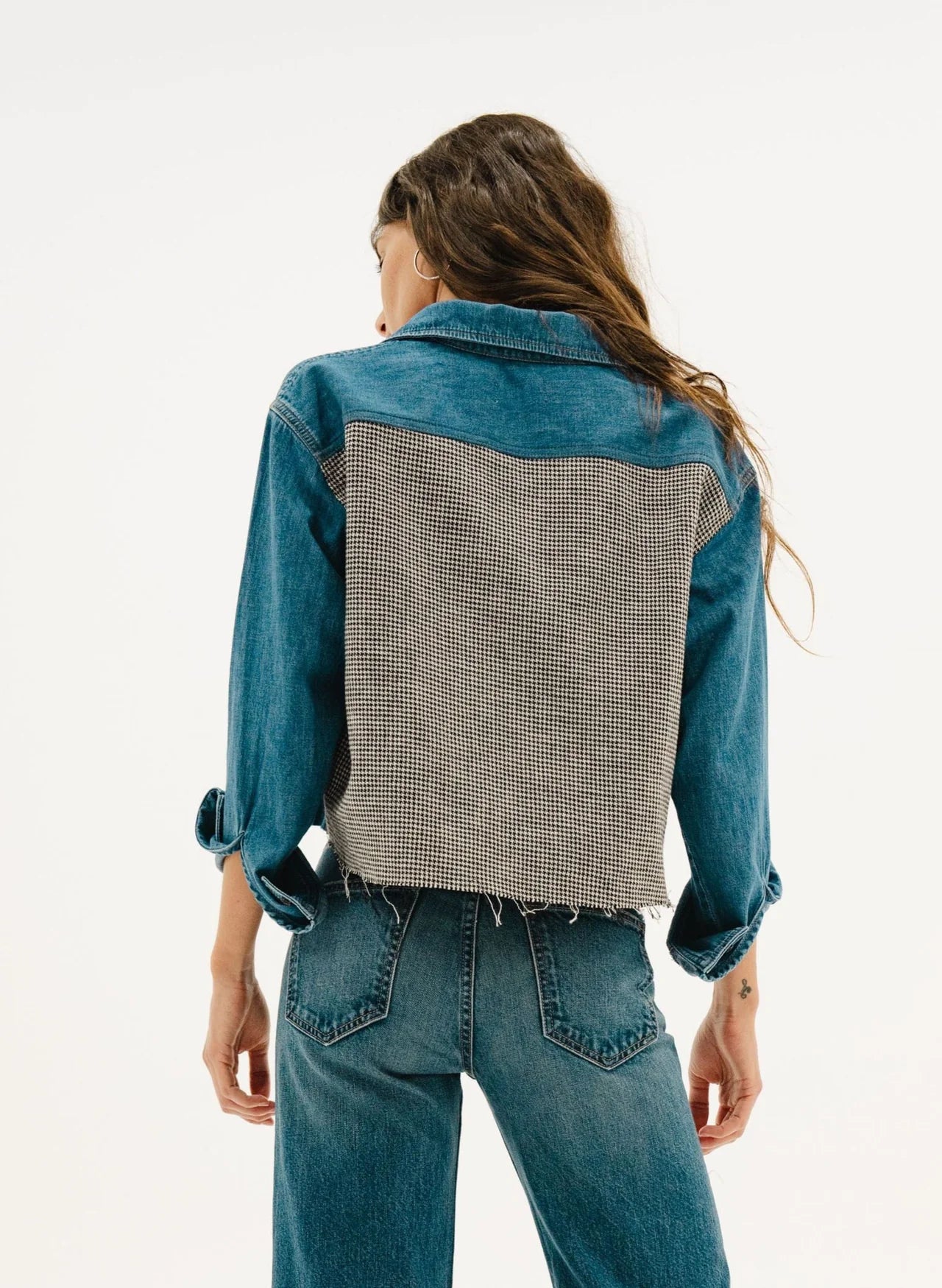A person with long wavy hair, facing away from the camera, wears the ASKK Crosby Flannel denim jacket with a Pink Plaid Japanese shirting fabric back and rolled-up sleeves, paired with blue jeans against a plain white background.
