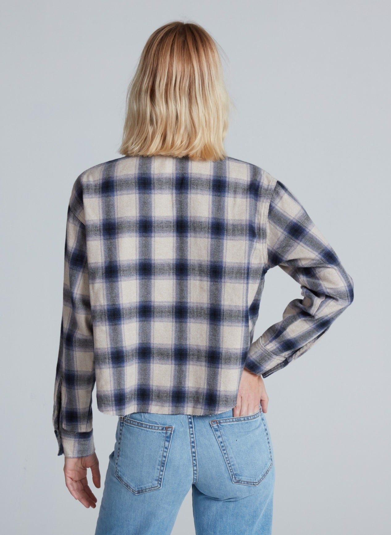 A person with shoulder-length blonde hair, seen from the back, wears the ASKK Crosby Flannel in blue and beige plaid with light blue jeans. Their left hand rests on their hip against a plain light gray background.