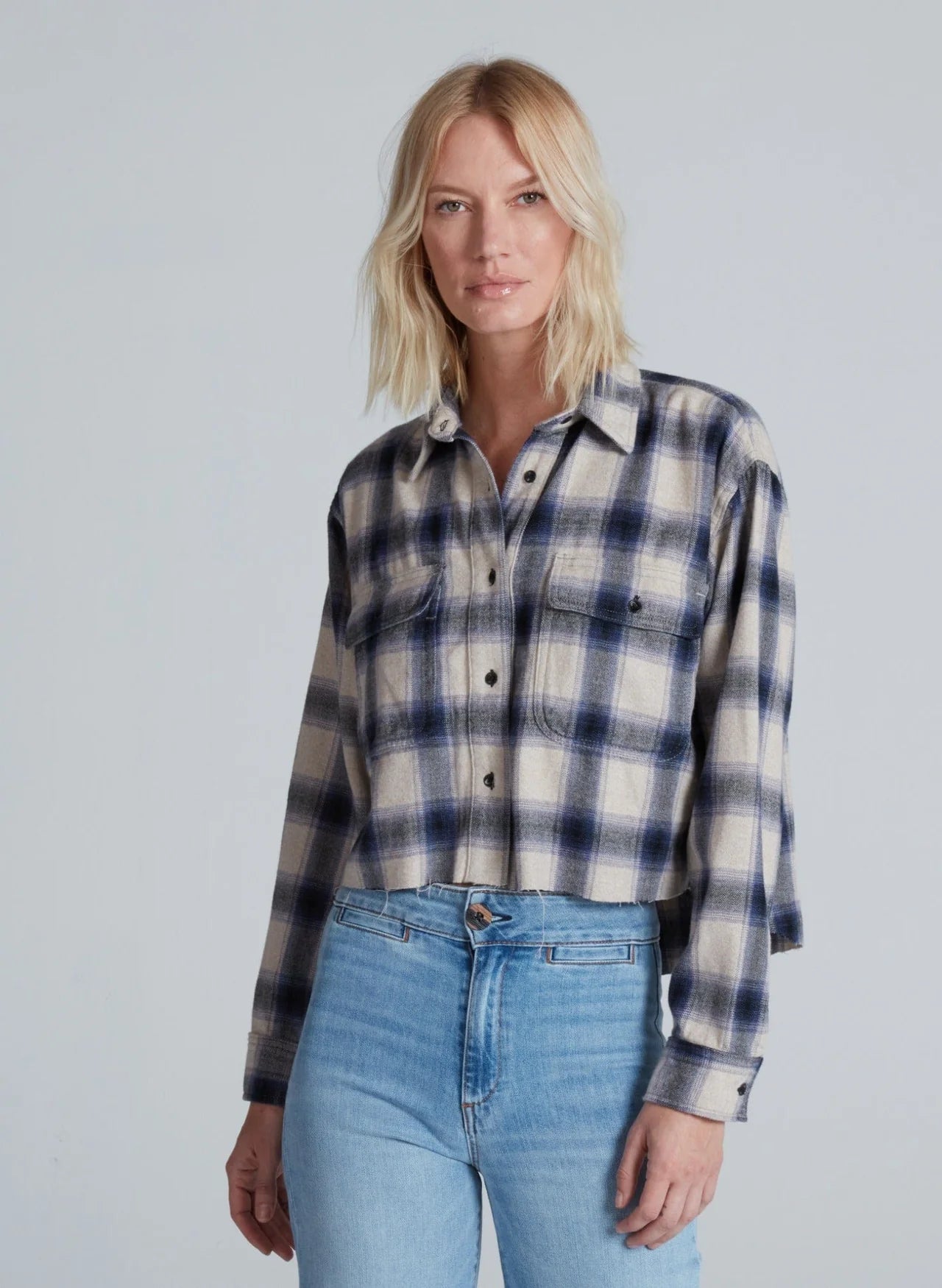 A woman with blonde, shoulder-length hair wears the ASKK Crosby Flannel in blue and white plaid, paired with light blue high-waisted jeans, standing against a plain light gray background.