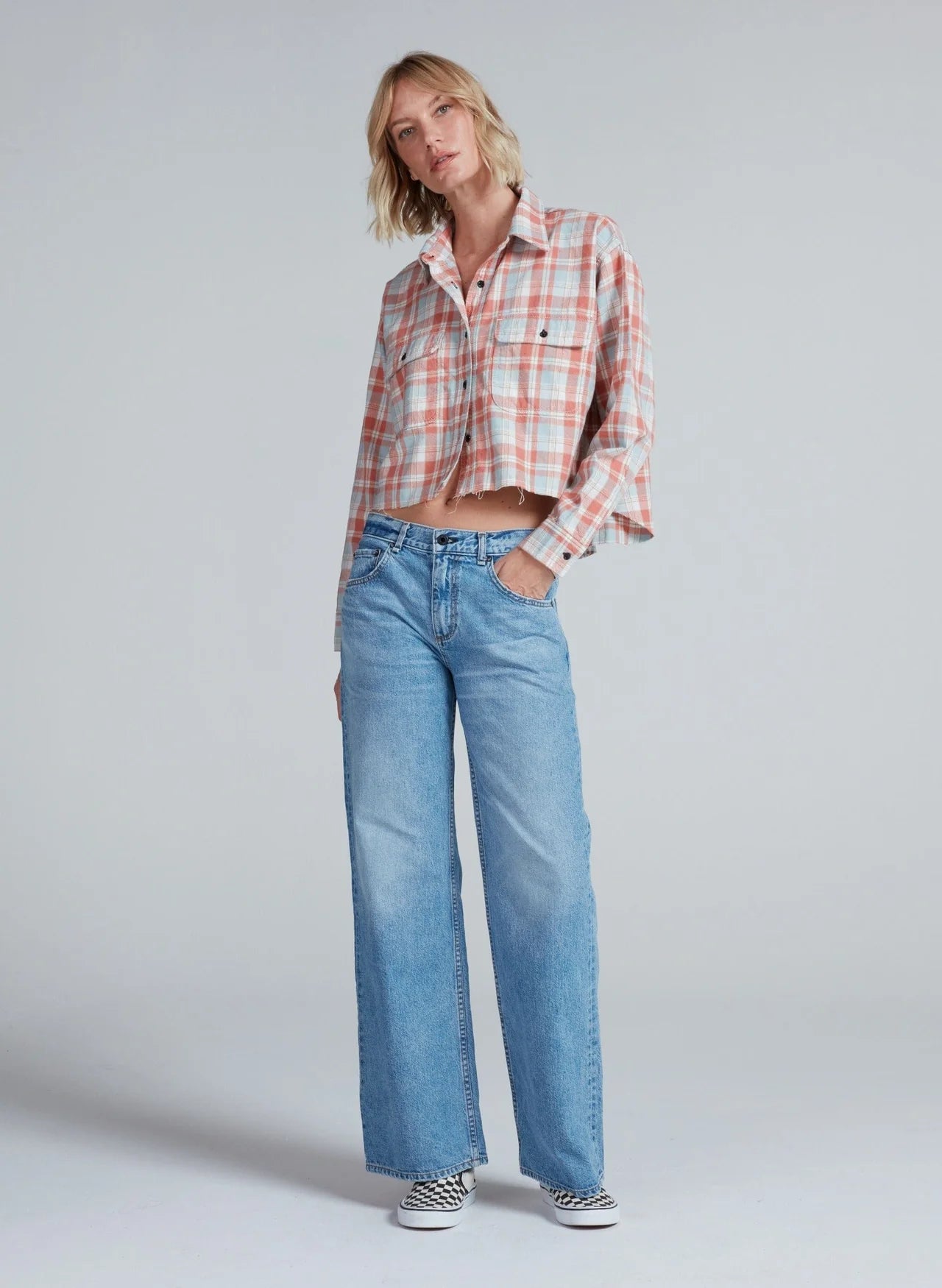 A person stands against a plain background wearing the ASKK Crosby Flannel—a cropped, long-sleeve plaid shirt—paired with light blue wide-leg jeans and checkered slip-on shoes. One hand is in their pocket, and they look relaxed.