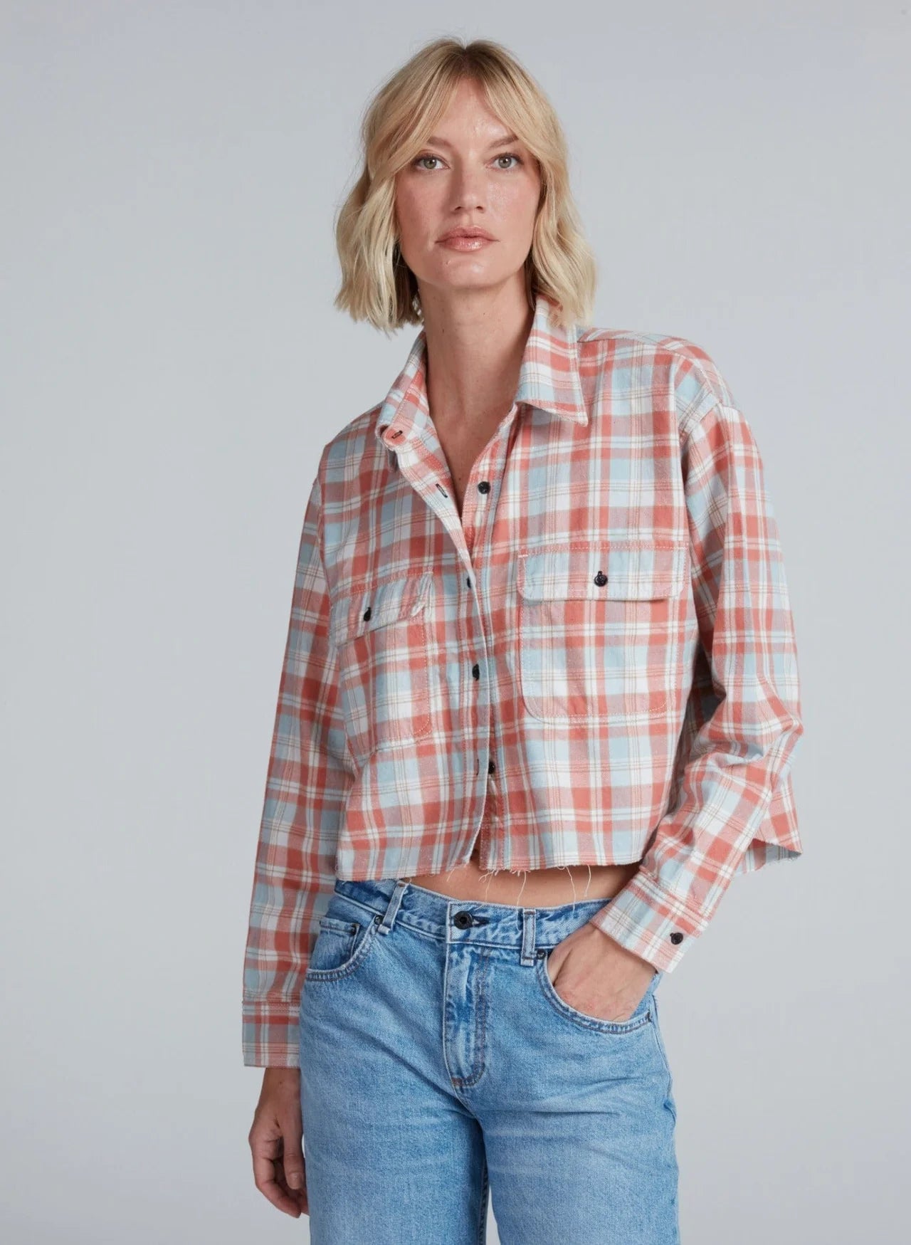 A woman with short blonde hair wears the ASKK Crosby Flannel—a cropped, long-sleeve plaid shirt in orange, blue, and white Japanese shirting—paired with light blue jeans. She stands against a plain background, one hand in her pocket.