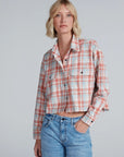 A woman with short blonde hair wears the ASKK Crosby Flannel—a cropped, long-sleeve plaid shirt in orange, blue, and white Japanese shirting—paired with light blue jeans. She stands against a plain background, one hand in her pocket.