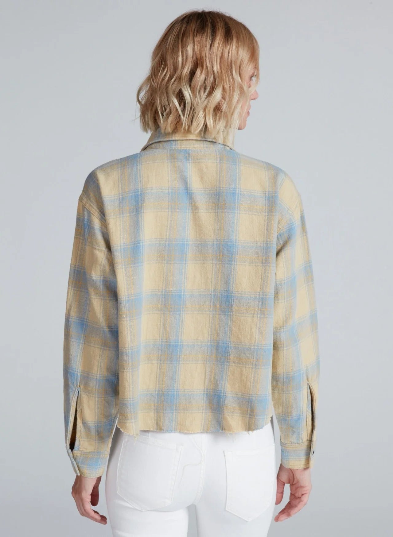 A person with short, wavy blonde hair is seen from the back in a beige and blue ASKK Crosby Flannel shirt made from Japanese shirting fabric, paired with white pants against a plain light gray background.