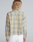 A person with short, wavy blonde hair is seen from the back in a beige and blue ASKK Crosby Flannel shirt made from Japanese shirting fabric, paired with white pants against a plain light gray background.