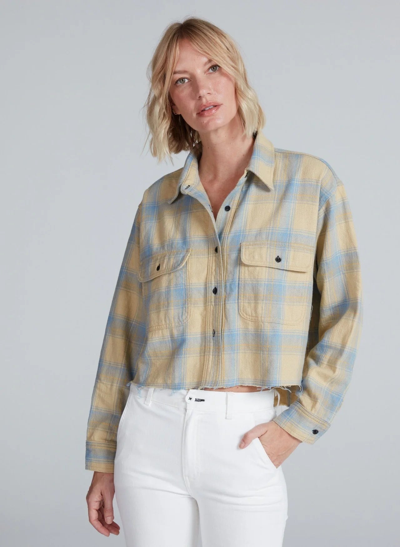 A woman with blonde, shoulder-length hair wears the ASKK Crosby Flannel, a beige and blue plaid shirt made from Japanese shirting fabric, paired with white high-waisted pants against a plain light gray background.