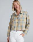 A woman with blonde, shoulder-length hair wears the ASKK Crosby Flannel, a beige and blue plaid shirt made from Japanese shirting fabric, paired with white high-waisted pants against a plain light gray background.