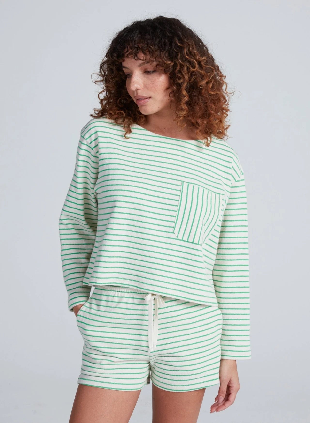 A person with curly hair wears the ASKK Easy Pull Over, a relaxed 100% cotton long-sleeve top with green stripes and a front pocket, paired with matching shorts, made in Los Angeles, against a plain light background.