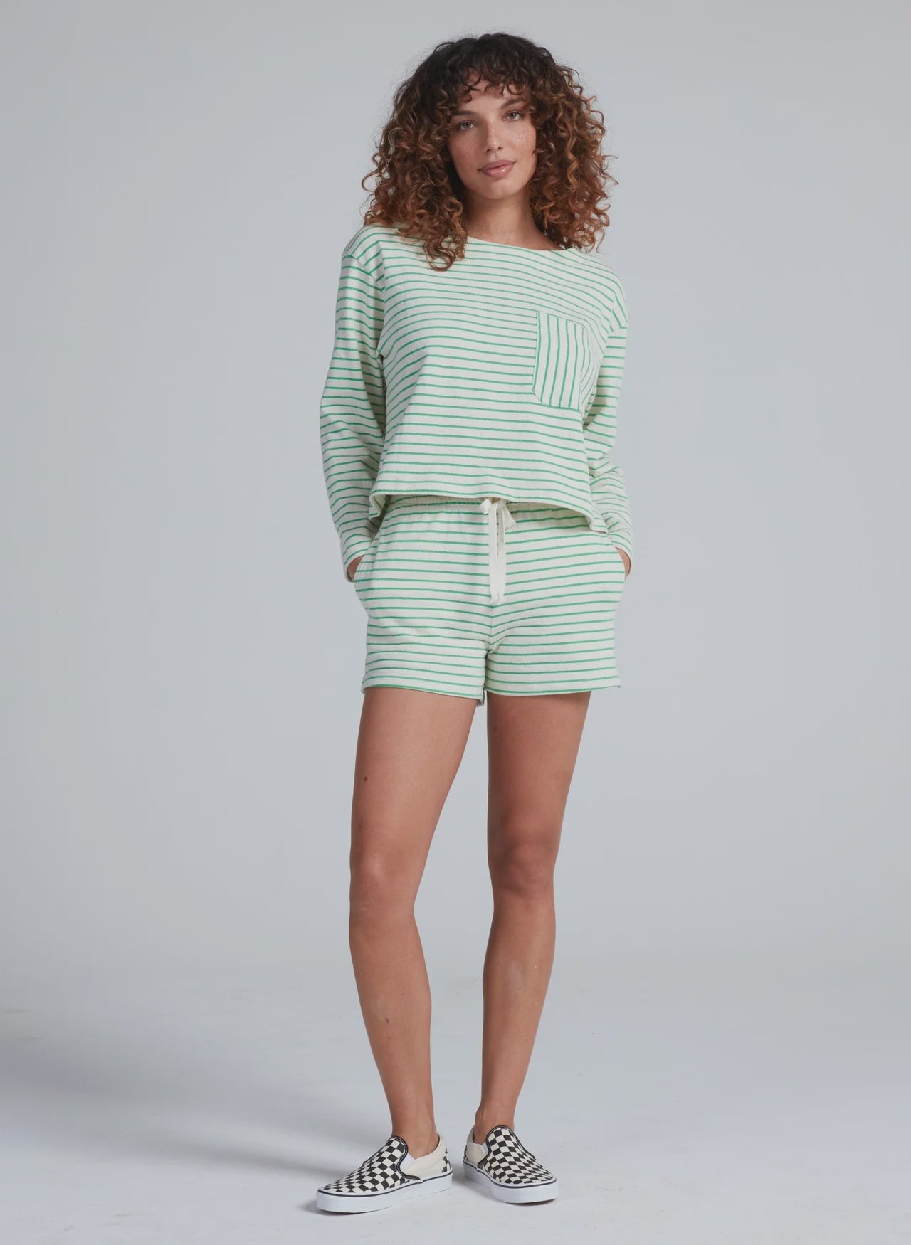 A person with curly hair wears the ASKK Easy Pull Over and matching light green and white striped shorts set, made in LA from 100% cotton, paired with checkered slip-on sneakers, standing against a plain light gray background.