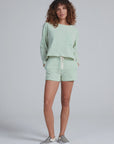 A person with curly hair wears the ASKK Easy Pull Over and matching light green and white striped shorts set, made in LA from 100% cotton, paired with checkered slip-on sneakers, standing against a plain light gray background.