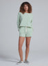 A woman poses casually against a plain light gray background, wearing ASKK’s Sweat Short paired with a mint green and white striped 100% cotton long-sleeve top with a pocket and checkered slip-on shoes.