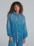 A woman with curly brown hair wears the ASKK Indigo Dip Dye Hoodie and matching joggers, featuring textured pockets and crafted from soft American knit cotton, standing against a plain light gray background.