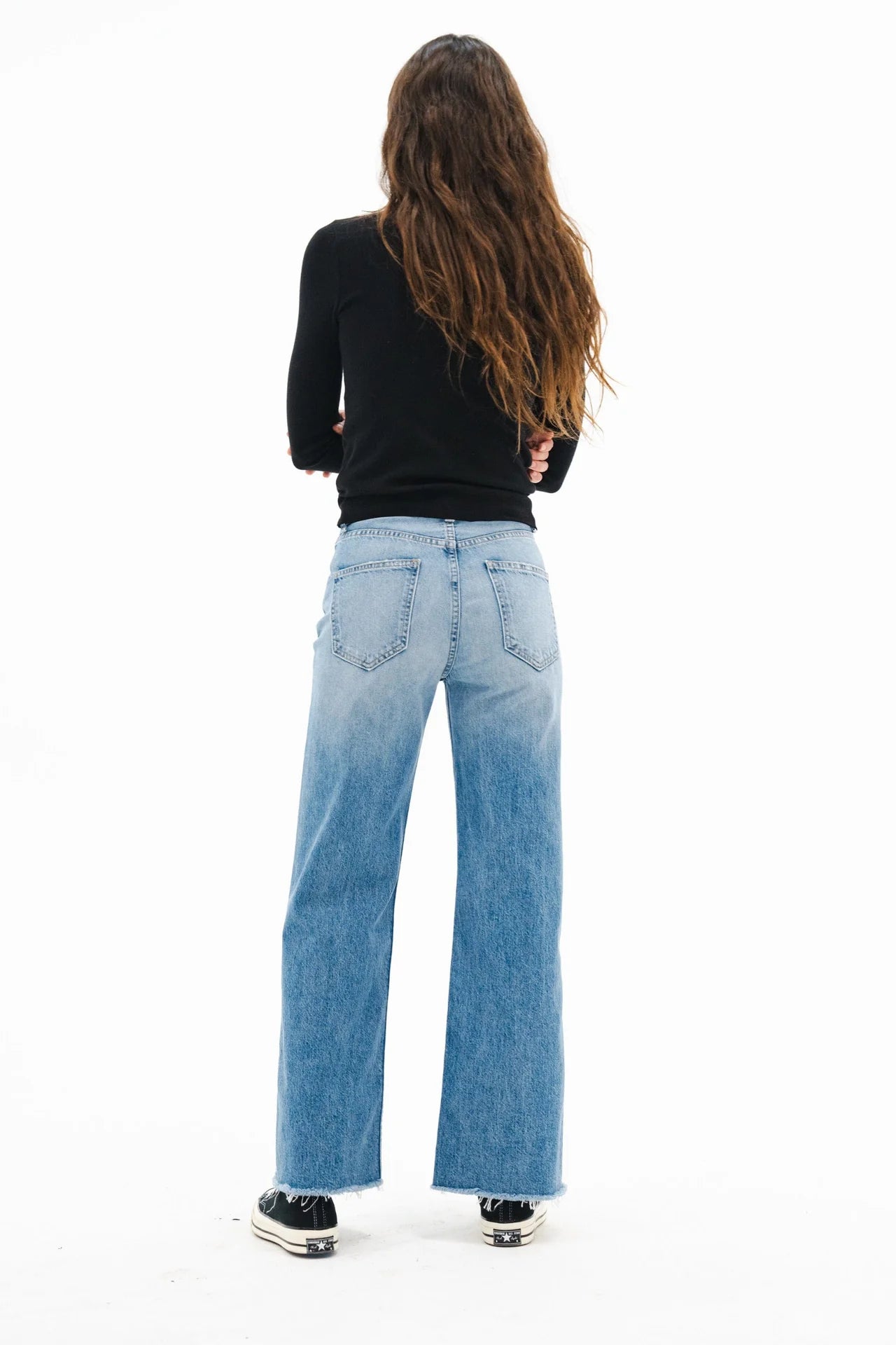 A person with long wavy hair, wearing a black long-sleeve top, ASKK Mercer Jean high-rise wide-leg denim, and black sneakers stands facing away from the camera against a white background.