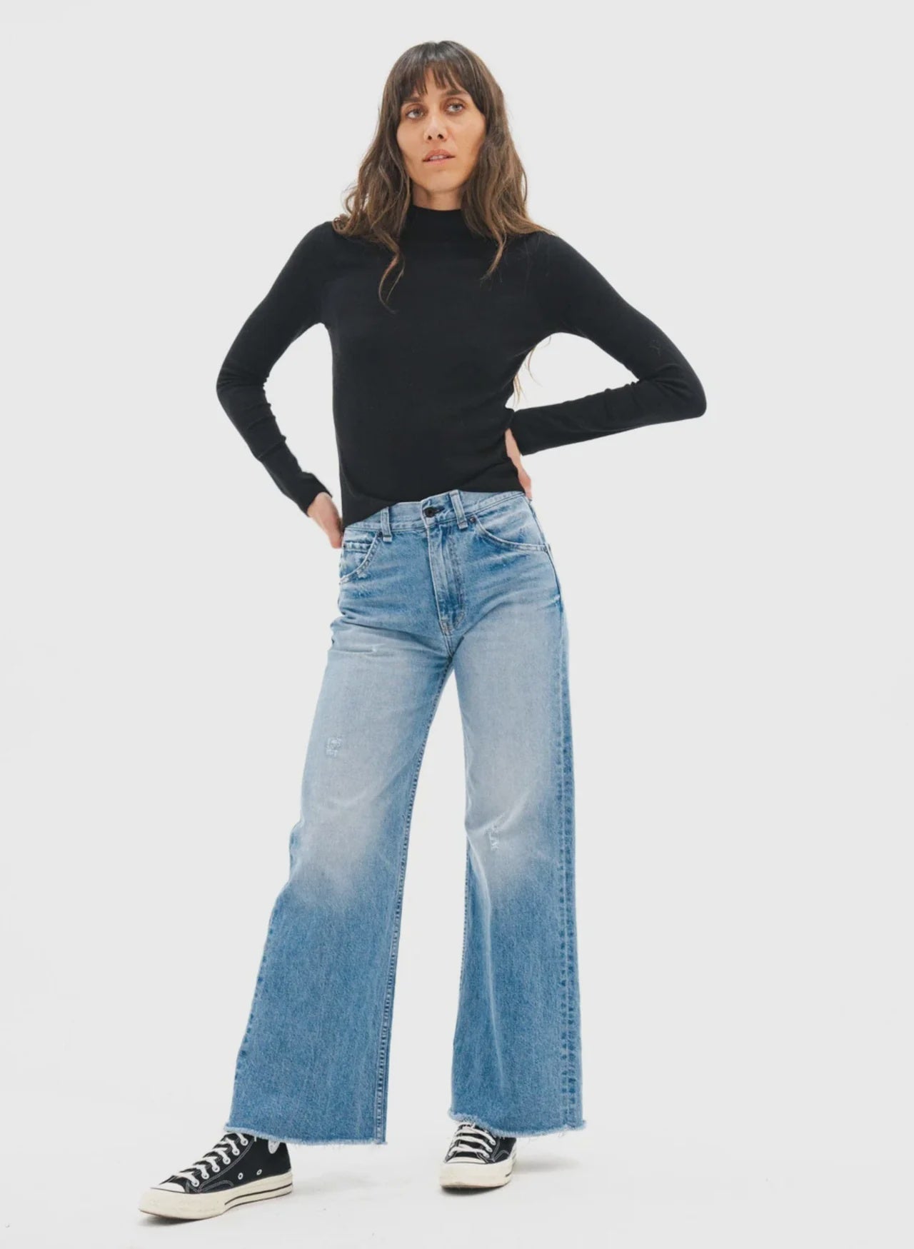 A woman poses against a plain background wearing a black long-sleeve turtleneck, ASKK Mercer Jean high-rise light blue wide-leg Japanese denim jeans, and black-and-white sneakers, her hands resting on her hips.