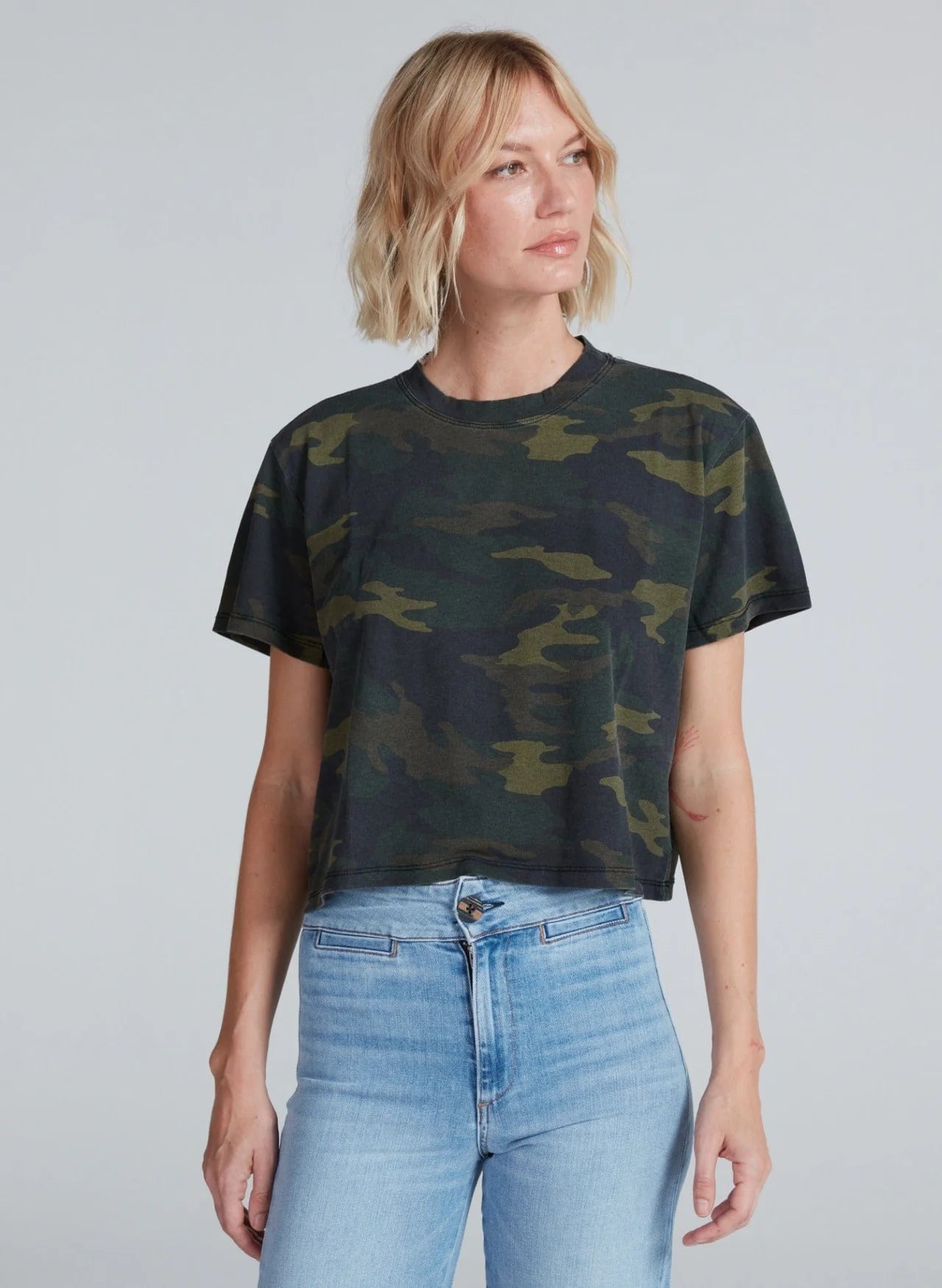 A person with short blonde hair wears the ASKK Shrunken Boy Tee in dark green camouflage and light blue high-waisted jeans, standing against a plain gray background and looking slightly to the side.