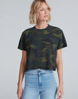 A person with short blonde hair wears the ASKK Shrunken Boy Tee in dark green camouflage and light blue high-waisted jeans, standing against a plain gray background and looking slightly to the side.
