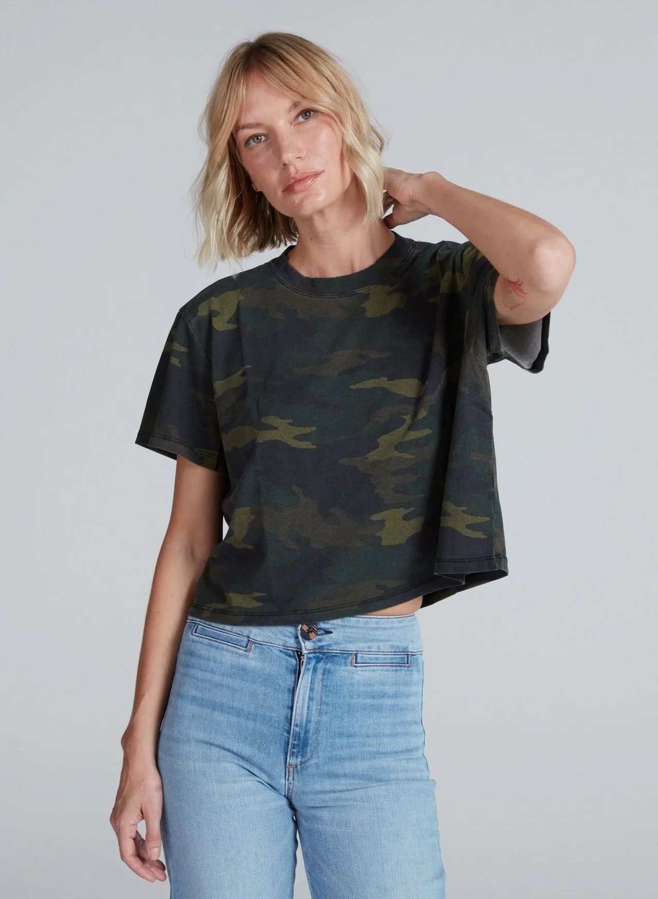 A woman with short blonde hair wears the ASKK Shrunken Boy Tee in dark camo print with light blue jeans, standing against a neutral background and resting one hand behind her neck.