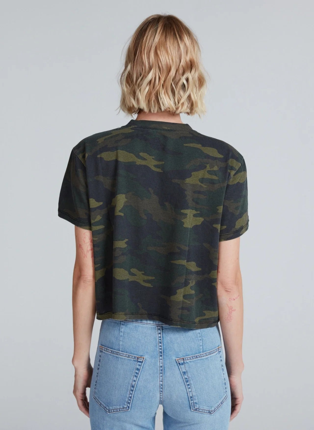 A person with short blonde hair is seen from the back, wearing the ASKK Shrunken Boy Tee in camouflage and high-waisted blue jeans against a plain light gray background.