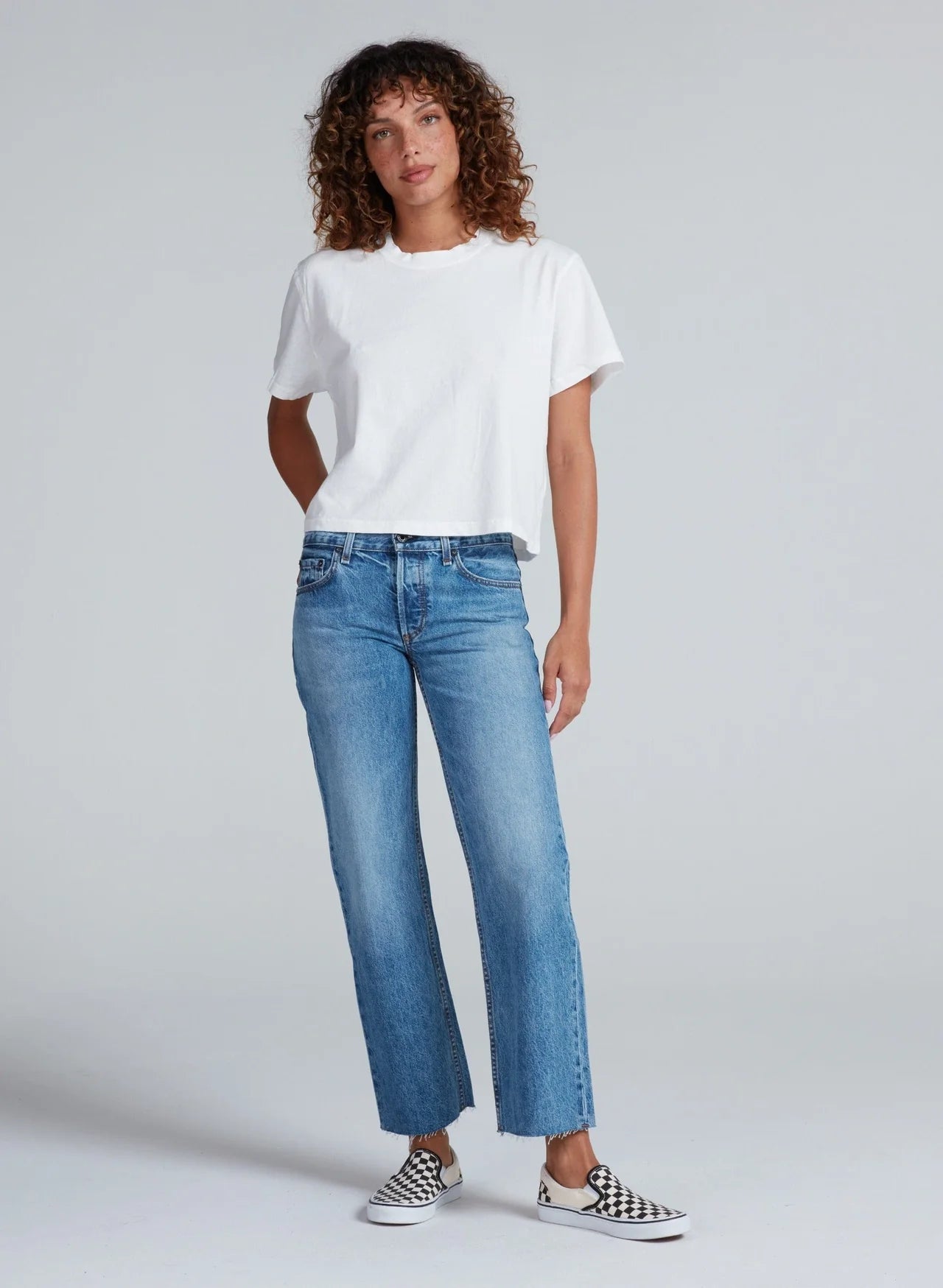 Someone with curly hair wears the ASKK Shrunken Boy Tee in white, blue straight-leg jeans, and black-and-white checkered slip-ons, standing against a plain light gray background.