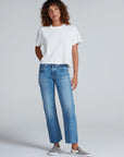 Someone with curly hair wears the ASKK Shrunken Boy Tee in white, blue straight-leg jeans, and black-and-white checkered slip-ons, standing against a plain light gray background.