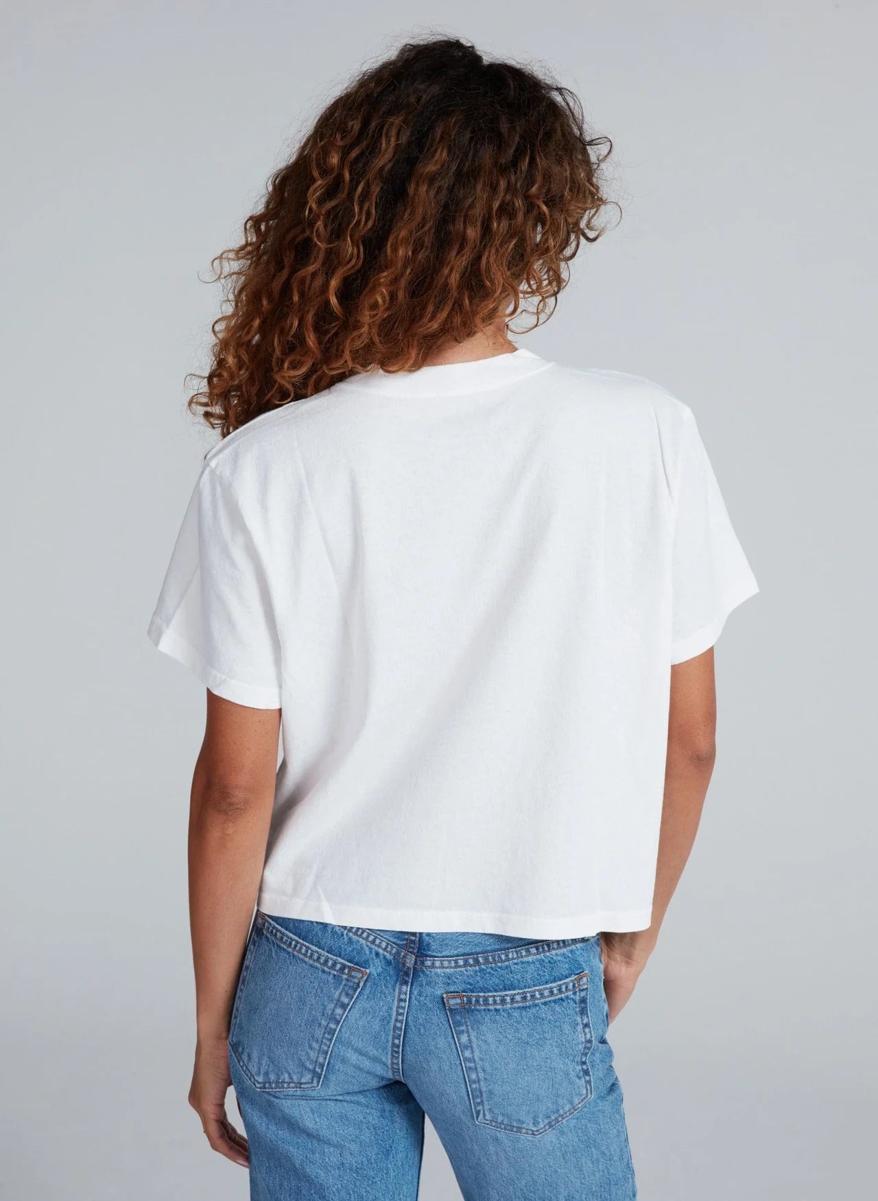 A person with curly brown hair is shown from the back, wearing an ASKK Shrunken Boy Tee and blue jeans against a light gray background.